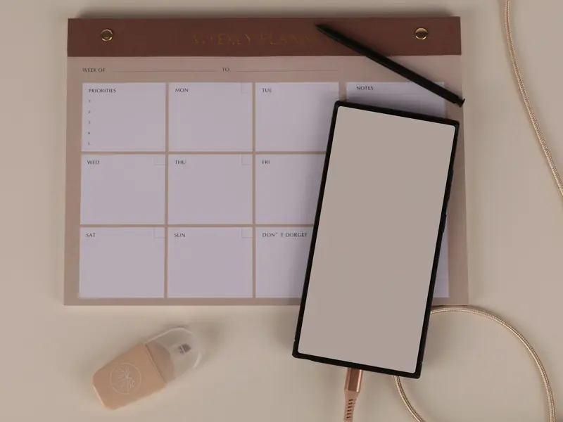 Blank weekly planner on a beige desk, with a smartphone and a pen.