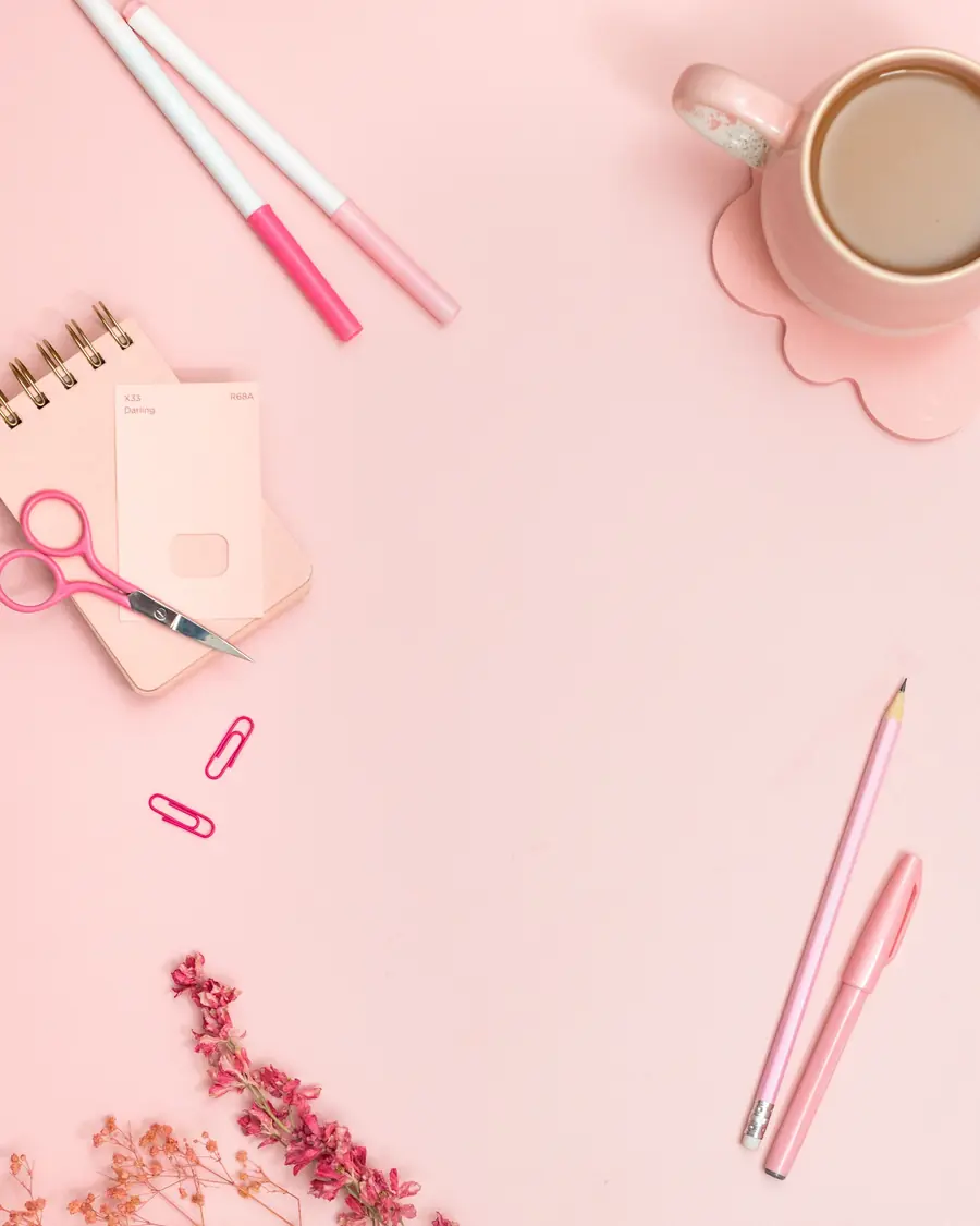 Pale pink desk seen from above: coffee cup, notebook, pens and dried flowers.